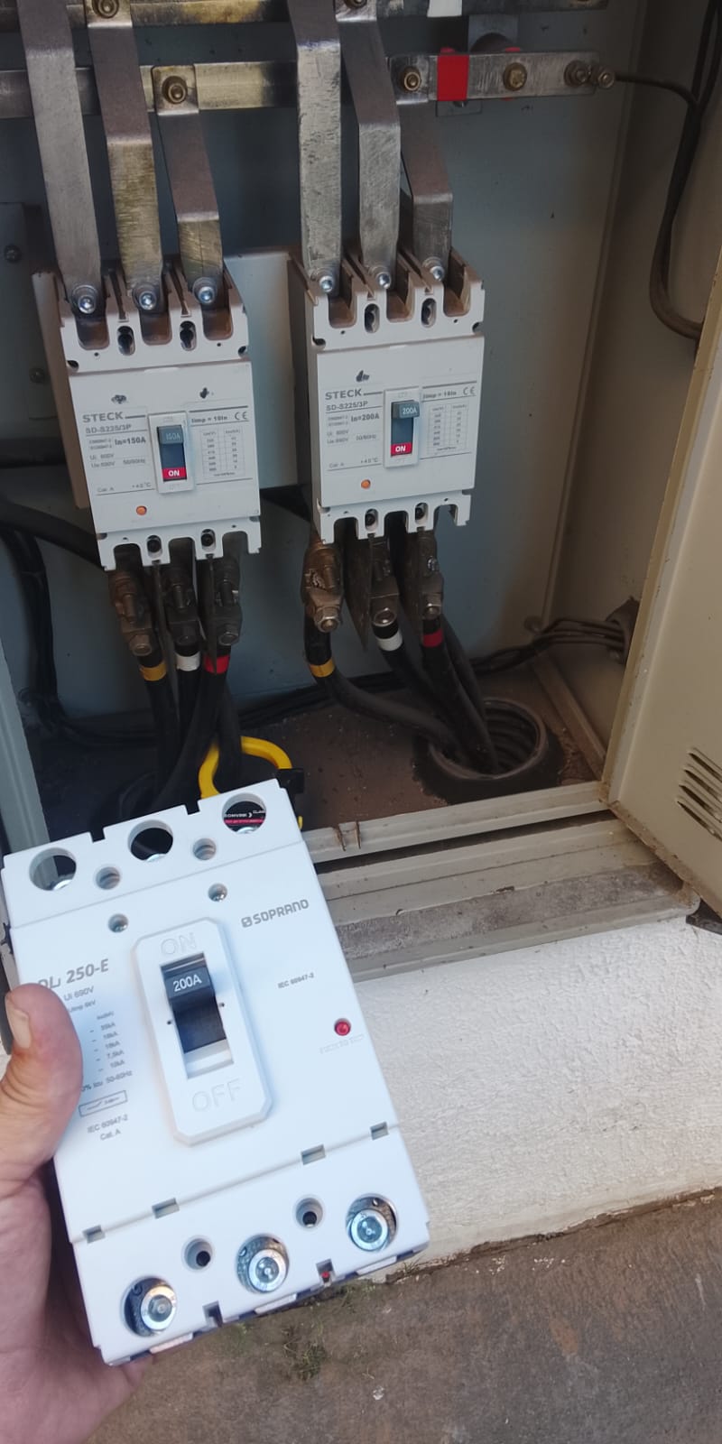 Painel elétrico com disjuntores Steck de 150A e 200A em manutenção, com novo disjuntor Soprano 200A em mãos para troca.